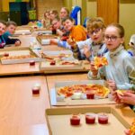 Dzieci siedzą przy stolikach i jedzą pizzę