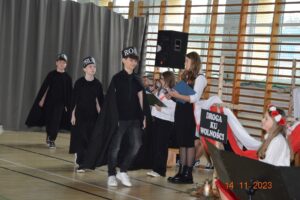 Chłopcy w czarnych pelerynach okrążają dziewczynkę, która gra Polskę