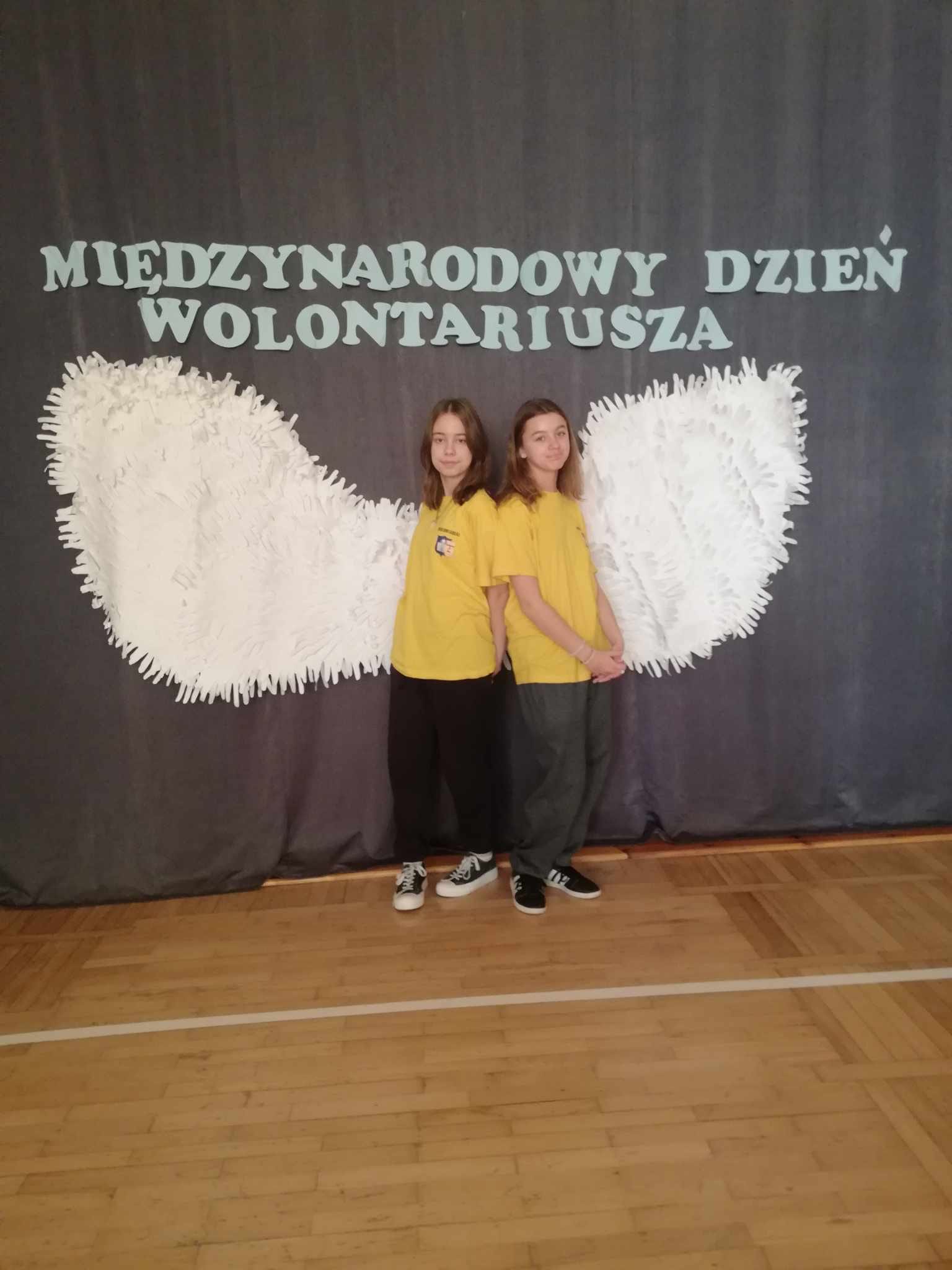 Dziewczynki na tle dekoracji- dużych białych skrzydeł i napisu Międzynarodowy Dzień Wolontariusza